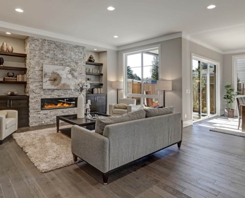 Living room interior in gray and brown colors features gray sofa atop dark hardwood floors facing stone fireplace with built-in shelves