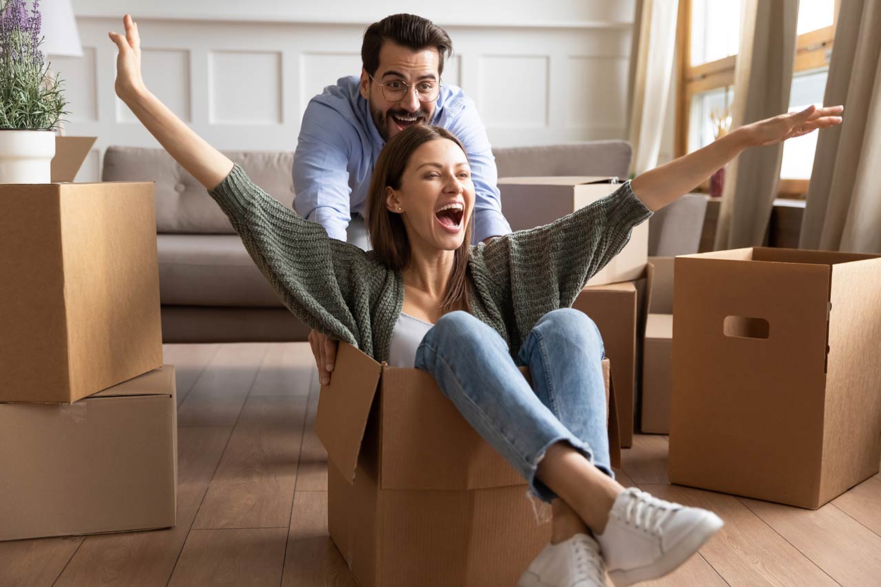 Happy family celebrating moving day, playful young couple having fun at home, funny activity, laughing husband pushing excited wife sitting in cardboard box