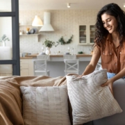 Lovely young woman putting soft pillows and plaid on comfy sofa, making her home cozy and warm